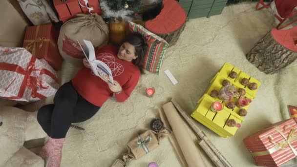 Έγκυος γυναίκα διαβάζει ένα βιβλίο κάτω από το χριστουγεννιάτικο δέντρο με λαμπερά χριστουγεννιάτικα φώτα - Πλάνα, βίντεο