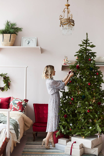 Beautiful and fashionable blonde model girl in trendy dress decorates Christmas tree in a bedroom. Young woman in modish outfit makes decoration in the interior for New year. Concept of Christmas eve - Фото, изображение