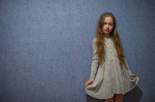 Schattig meisje van elf jaar oud met blond lang haar in een grijze jurk op een blauwe achtergrond - Foto, afbeelding