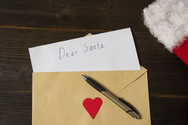 Письмо Санте в конверте. Ручка и шляпа на деревянном фоне. Концепция Рождества
 - Фото, изображение