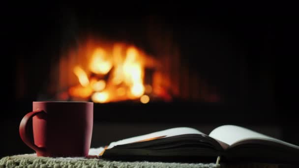 Otevřená kniha a šálek horkého čaje na pozadí krbu, kde hoří oheň - Záběry, video