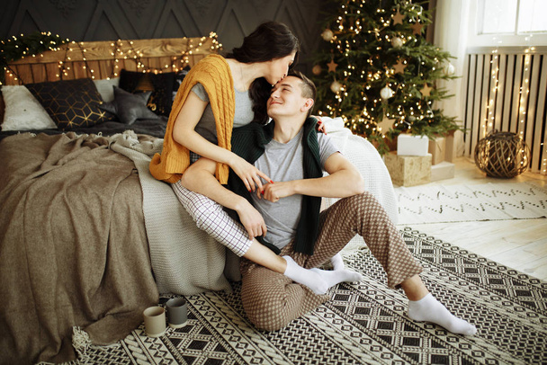 happy couple in love in the morning on the bed near the Christmas tree, hug, Christmas morning. home clothes and sweaters. comfort and warmth of hearts - Valokuva, kuva