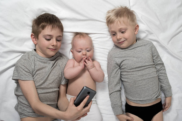 Deux frères avec un plus jeune enfant sont allongés sur un lit blanc et prennent du selfie sur un smartphone. Bonne enfance, grande famille
 - Photo, image