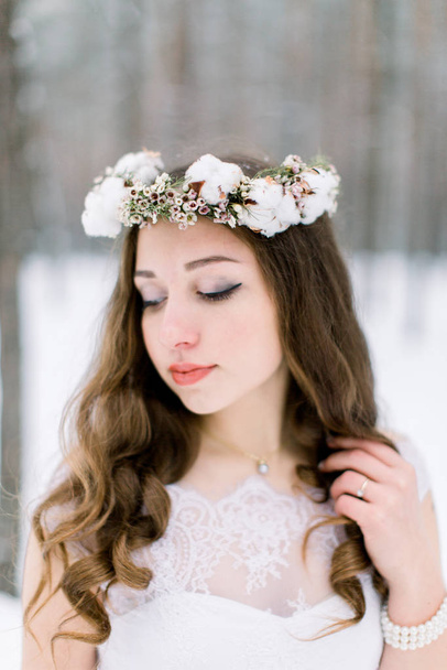 Beautiful bride touching her curly hair, with floral head band wreath, made from flowers and cotton, standing outdoors, on the background of winter forest - Foto, Imagem