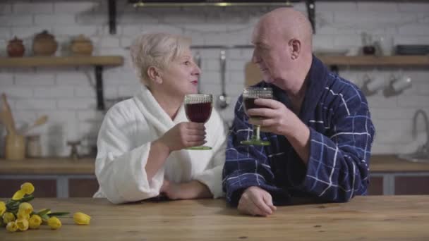 Smiling senior Caucasian husband and wife sitting at the table with wineglasses and talking. Old spouses in bathrobes resting in the evening at home. Eternal love, happiness, togetherness. - Materiał filmowy, wideo