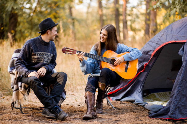 Portrait of young couple sitting on camp chair with guitar near camp tent. Time together in harmony with nature far from the bustle of the city - Foto, afbeelding