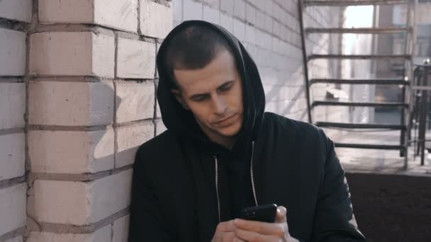 Retrato de joven gangster traficante de drogas en el capó utiliza un teléfono inteligente fondo urbano al aire libre
 - Metraje, vídeo