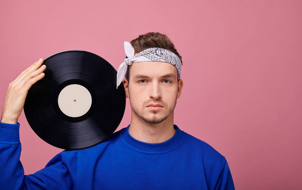 The average plan. A cool stylish guy in a bandana in a dark blue jacket stands on a pink background with a black vinyl record in his hand. Youth, style, confidence, joy, playful mood. Looking straight - Foto, Imagem