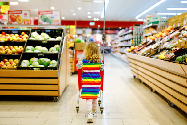 Cute todler girl pushing shopping cart in supermarket. Little child buying fruits. Kid grocery shopping. Adorable baby kid with trolley choosing fresh vegetables in local store. - Photo, Image