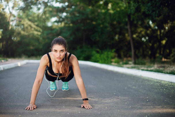 Alluring determined young sporty woman in activewear standing in plank concrete road in park, perform push-up exercises during daily morning warm-up before jogging, listen music wired earphones - Photo, Image