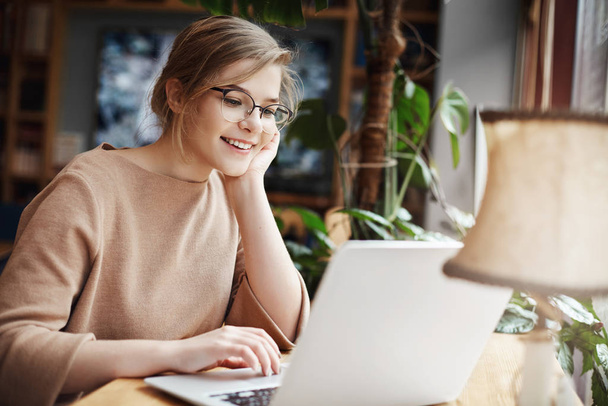 Cheerful young caucasian girl in glasses using laptop, shopping online or browsing internet, chatting with friends, edit photos for university project, smiling as looking computer screen - Fotó, kép