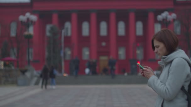 Young woman looking at her smartphone with red building behind. Female tourist in gray coat with backpack walking near city sight university.  - Felvétel, videó