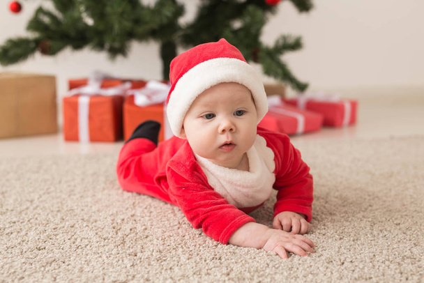 Χαριτωμένο κοριτσάκι που φοράει κοστούμι Άη Βασίλη σέρνεται στο πάτωμα πάνω από το χριστουγεννιάτικο δέντρο. Περίοδος διακοπών. - Φωτογραφία, εικόνα