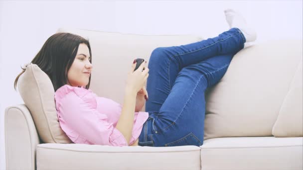 Cute caucasian girl is lying on the sofa, putting legs on the back of the sofa, using phone, messaging. - Imágenes, Vídeo