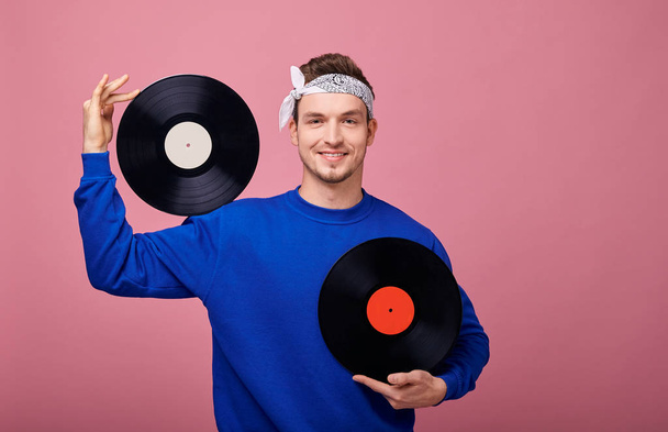 Retro style smiling hippie man in white bandana in a blue sweater holds one black vinyl on the shoulder, and holds the second record in left hand at chest level. Close-up. Happy, joyful. - Foto, Imagen
