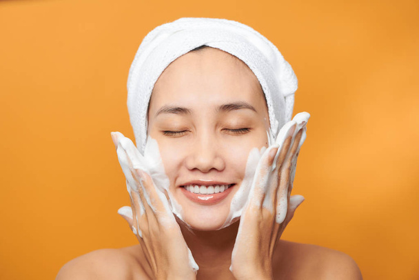 Счастливая молодая азиатка наносит крем для лица, надевая полотенце и трогая лицо. Изолированный на оранжевом фоне
 - Фото, изображение