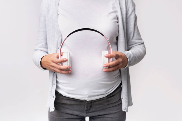 Неузнаваемая беременная женщина, держащая наушники возле живота на сером фоне
 - Фото, изображение
