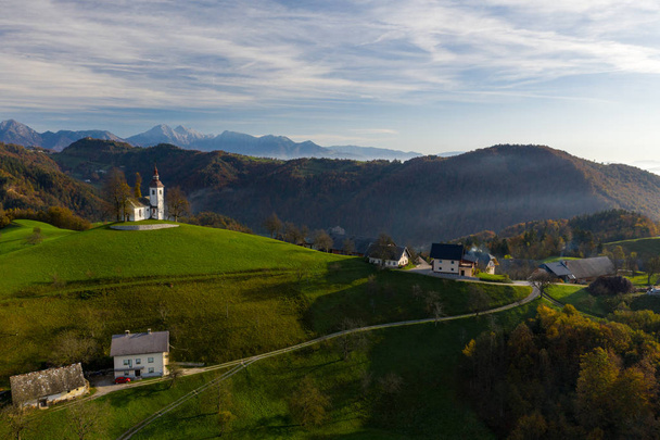 Aerial view of the hills, colorful forest in the fog and the church of Sv Tomaz. Sunrise in Slovenia in autumn - Photo, Image