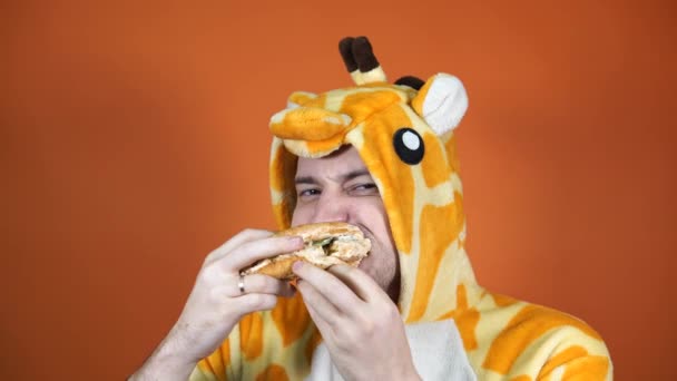 スタジオのオレンジの背景にハンバーガーを食べているキリンの大きなパジャマのハンサムな男. - 映像、動画
