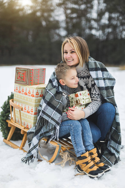 Mother with little son in winter park sledging. Mom and child sitting together on wooden decorated sledge with Christmas presents. Winter walk outdoors - Фото, изображение