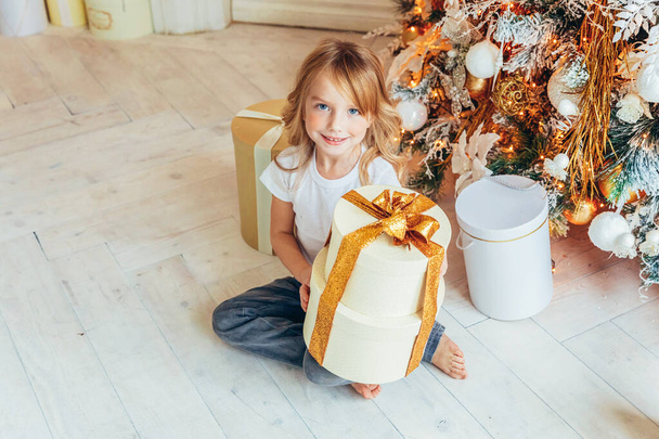 Little girl with gift box near Christmas tree on Christmas eve at home. Young kid in light room with winter decoration. Happy family at home. Christmas New Year december time for celebration concept - Photo, Image