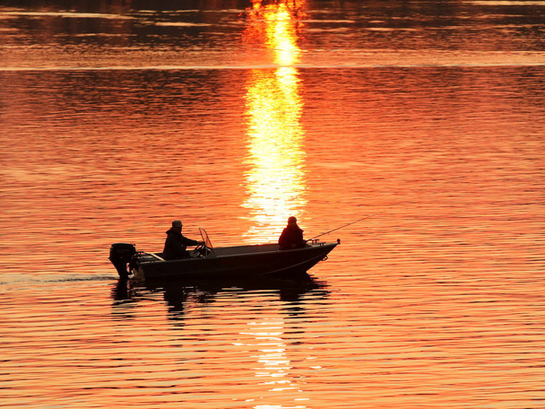 Dwóch rybaków, mężczyzna i kobieta, pływają w motorówce na rzece na tle pomarańczowego zachodu słońca. Romantyczna atmosfera dla kochanków. Wieczorny połów. Podróż przez wodę. Naturalne zjawisko - Zdjęcie, obraz