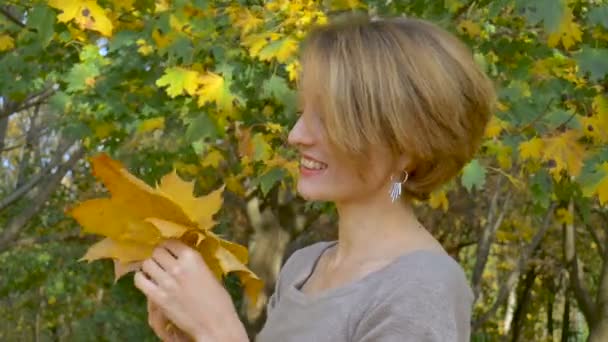Emotional attractive young woman with blonde short hair and biege dress holding a bouquet of autumn leaves above her head and posing in a beautiful park outdoors - Video, Çekim