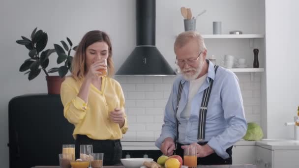 cute elderly man with a beard in glasses for vision has fun with granddaughter while preparing lunch from healthy products, drinking juice and chatting in kitchen at table - Video