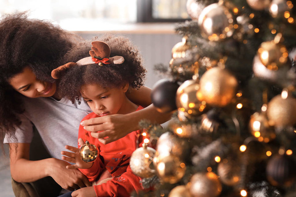 Happy African-American woman and her little daughter decorating Christmas tree at home - 写真・画像