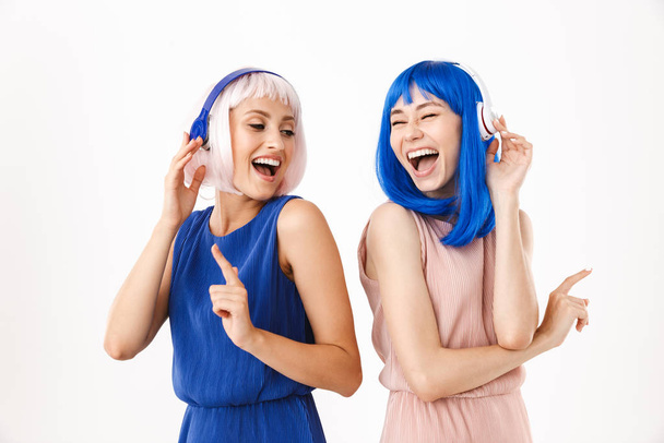 Portrait of two joyful women wearing blue and pink wigs pointing fingers aside while using headphones - Photo, Image