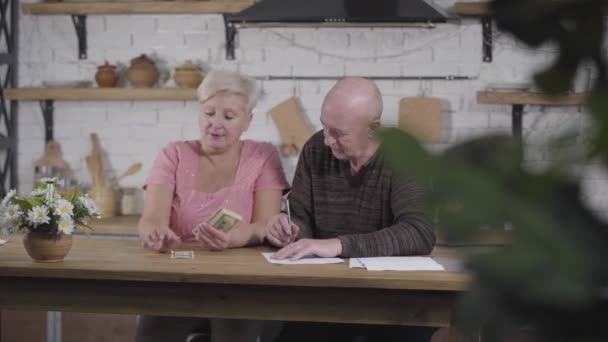 Mature Caucasian husband and wife settling their income and expenses. Woman counting cash, man writing down budget. Old spouses settling their financials. - Πλάνα, βίντεο