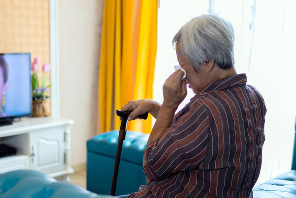 Ασιάτισσα ηλικιωμένη γυναίκα κλαίει, κρατά ένα χαρτί υγείας στο χέρι και σκουπίζοντας στα μάτια της μορφή βλέποντας ταινίες σειρές δράματα στην τηλεόραση, ηλικιωμένοι απολαμβάνουν να παρακολουθήσετε λυπητερή ταινία σε μια καθημερινή ρουτίνα ή εθισμένοι στο δράμα, τον τρόπο ζωής - Φωτογραφία, εικόνα