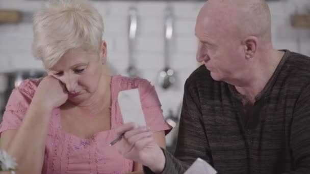 Close-up of serious mature Caucasian man showing bills to his ashamed wife. Beautiful senior lady closing eyes with hand and taking papers from her husband. Wife spending family budget, retirement. - Кадри, відео