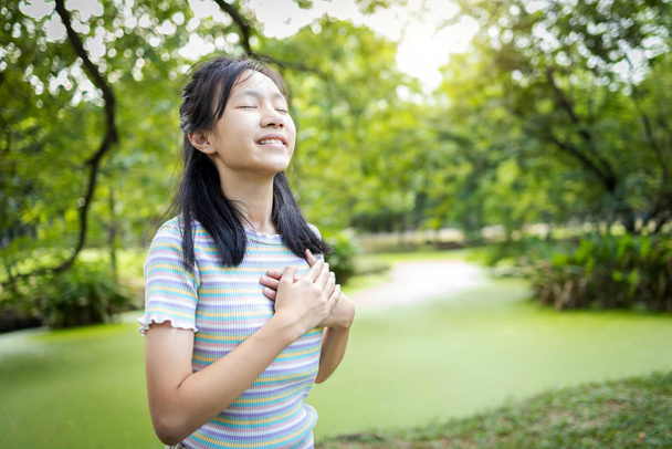 Gelukkig glimlachend aziatisch kind meisje staan in de groene natuur, vasthouden ands op hart, vrouwelijke tiener aanraken van haar borst, genieten van frisse lucht met gesloten ogen, ontspannen in het park, gezonde levensstijl concept - Foto, afbeelding