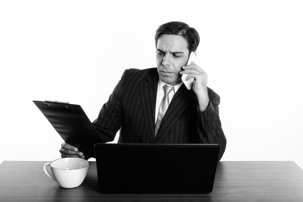 Uomo d'affari persiano che parla sul cellulare mentre legge negli appunti con laptop e tazza di caffè sul tavolo di legno
 - Foto, immagini