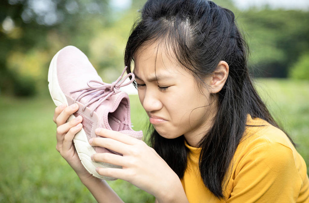 unglückliche asiatische Teenagerin schnüffelt an ihren Turnschuhen, hält mit Ekel Stinkehacke in der Hand, unangenehmer Geruch wegen des heißen Wetters oder nach dem Sport, trauriges Kindermädchen mit schlechtem Geruch, angehäufter Schmutz - Foto, Bild