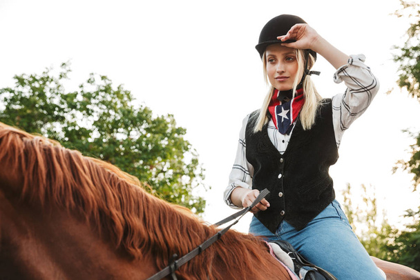 Image of adorable woman wearing hat riding horse at yard in coun - Photo, Image