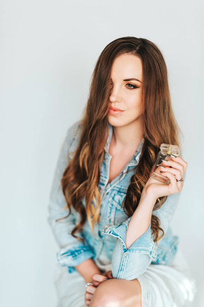 Egy fiatal, gyönyörű nő portréja hosszú frizurával, kék farmerdzsekiben, kezében egy üveg parfümmel. - Fotó, kép