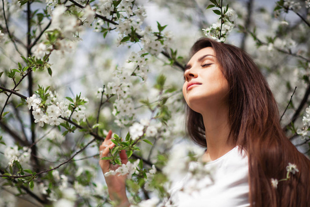 Girl standing under blossoming apple tree in natural park. Fashion concept. Elegant young lady in jeans clothes enjoying flowering garden at sunny spring day.Natural beauty makeup. Horizontal - Photo, Image