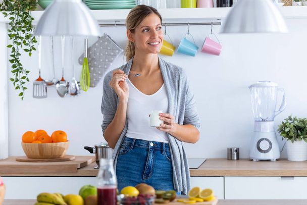 Гарненька молода жінка їсть йогурт, стоячи на кухні вдома
. - Фото, зображення