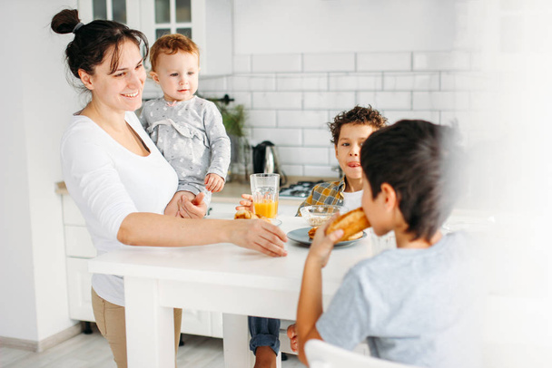 Молодая женщина мама с ребенком девочка на руках готовить завтрак на яркой кухне дома, большая счастливая семья
 - Фото, изображение