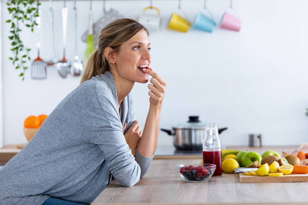 Kaunis nuori nainen syö punaisia marjoja istuessaan keittiössä
 - Valokuva, kuva