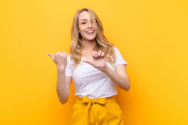 young pretty blonde woman smiling cheerfully and casually pointing to copy space on the side, feeling happy and satisfied against flat color wall - Foto, imagen