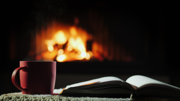 Otwarta książka i filiżanka gorącej herbaty na tle kominka, gdzie pali się ogień. - Zdjęcie, obraz
