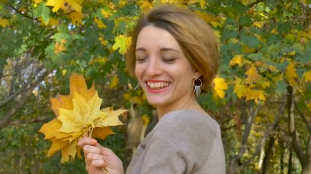Emotional attractive young woman with blonde short hair and biege dress holding a bouquet of autumn leaves above her head and posing in a beautiful park outdoors - 映像、動画