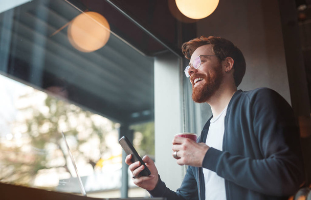 Cheerful bearded male with coffee and smartphone - Zdjęcie, obraz
