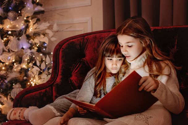 Szczęśliwe młode siostry czytające razem książkę do opowiadania przy kominku w przytulnym ciemnym salonie w Wigilię. Świętowanie Bożego Narodzenia w domu. - Zdjęcie, obraz