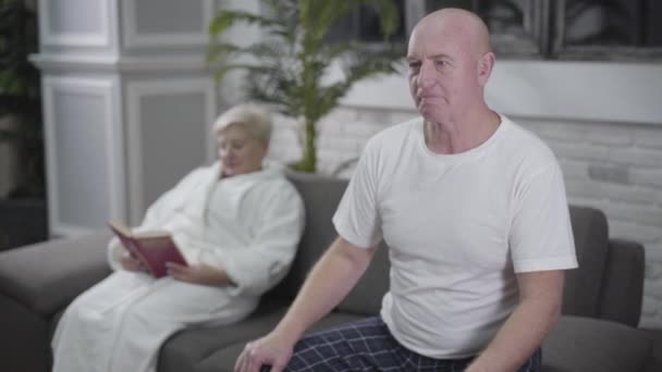 Concentrated senior bald man sitting on couch and watching TV when suddenly having heartache. His blond Caucasian wife putting aside book and coming to husband from background. Medicine, health, care. - Кадри, відео