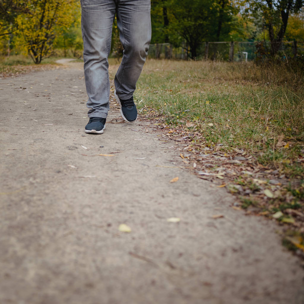 Piernas masculinas en jeans y zapatillas de deporte caminando hacia la distancia en el suelo cubiertas de hojas amarillas caídas. Concepto de llegada de otoño, primer plano, espacio para copiar
. - Foto, imagen