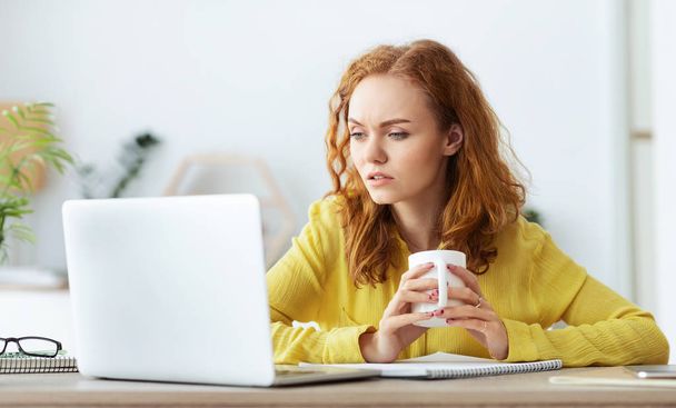 Σοβαρό κορίτσι διαβάζει ειδήσεις στο laptop και πίνοντας καφέ - Φωτογραφία, εικόνα